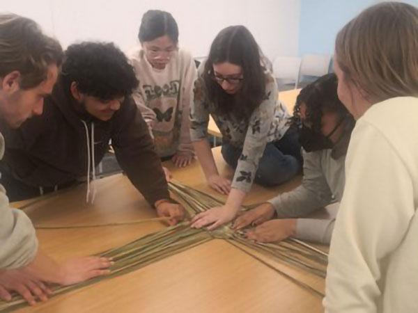 Students gather around a table and work together to weave a square using natural materials